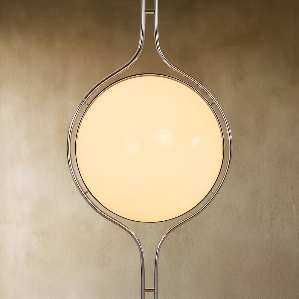 Clérachic floor-to-ceiling lamp in silver, the globe lit warm and centred in its ring against a stone-coloured wall.