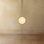 Clérachic floor-to-ceiling lamp in brass, the lit globe on a plain plaster wall and the twin-rod pole running down to a small round foot on a stone floor.