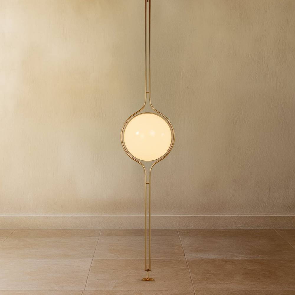 Clérachic floor-to-ceiling lamp in brass, the lit globe on a plain plaster wall and the twin-rod pole running down to a small round foot on a stone floor.