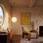 Clérachic floor-to-ceiling lamp in brass, standing between a round window and a plaid armchair, its pole rising to a small round ceiling canopy.