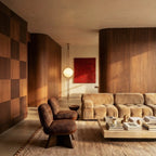 Clérachic floor-to-ceiling lamp in brass, lit in a wood-panelled living room beside a red painting and a tan sectional sofa.