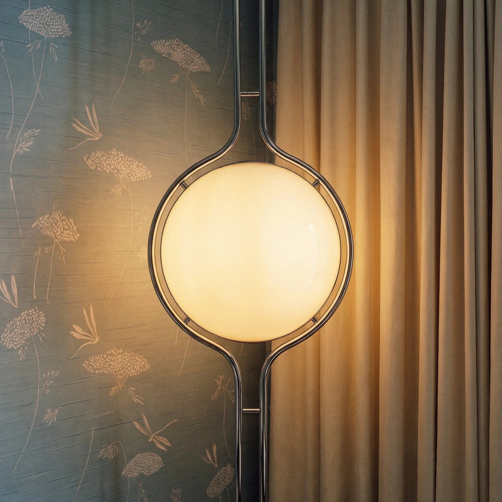 Clérachic floor-to-ceiling lamp lit against grey-blue wallpaper printed with pale flowers, its polished ring catching a sharp highlight beside a curtain.