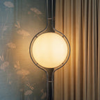 Clérachic floor-to-ceiling lamp lit against grey-blue wallpaper printed with pale flowers, its polished ring catching a sharp highlight beside a curtain.