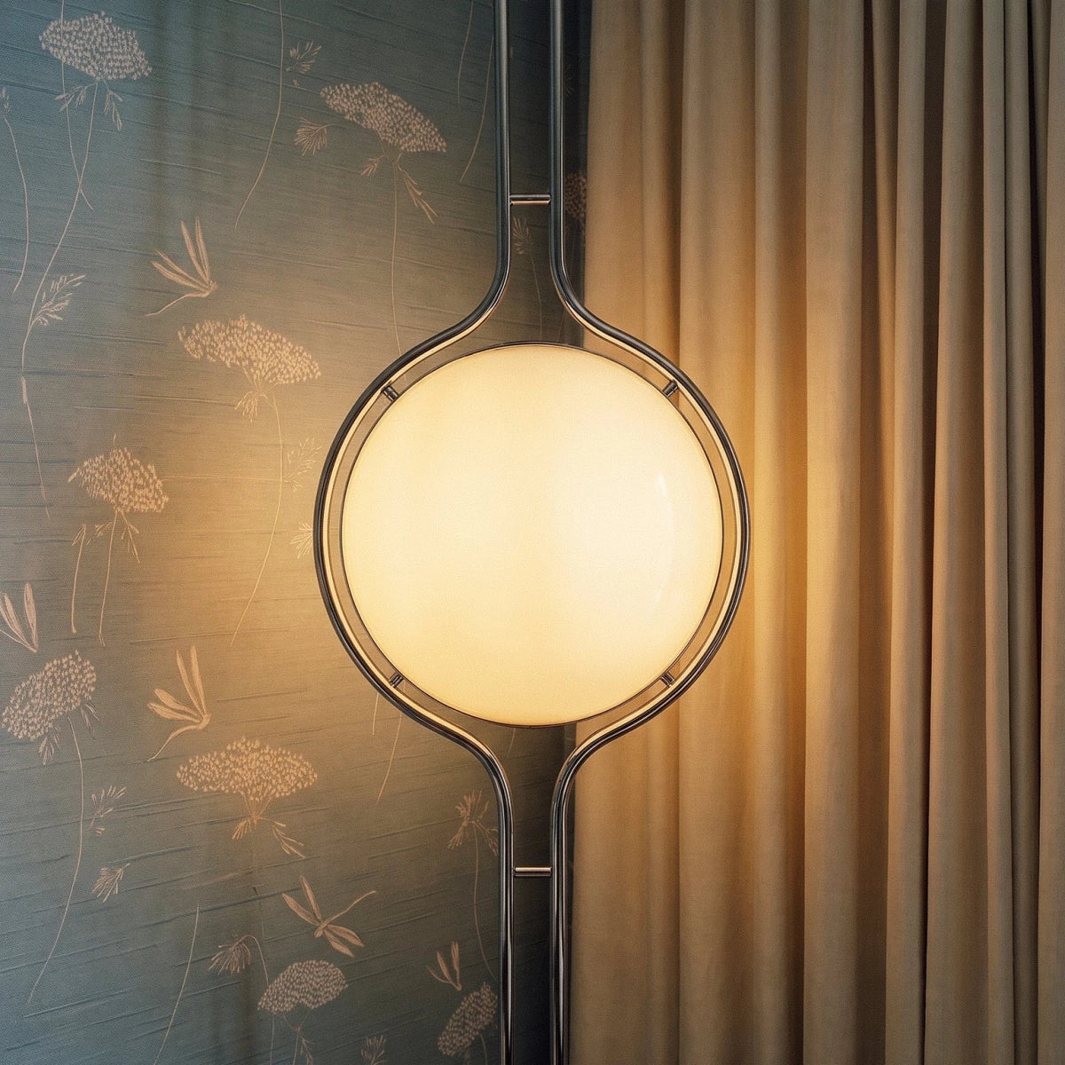 Clérachic floor-to-ceiling lamp lit against grey-blue wallpaper printed with pale flowers, its polished ring catching a sharp highlight beside a curtain.