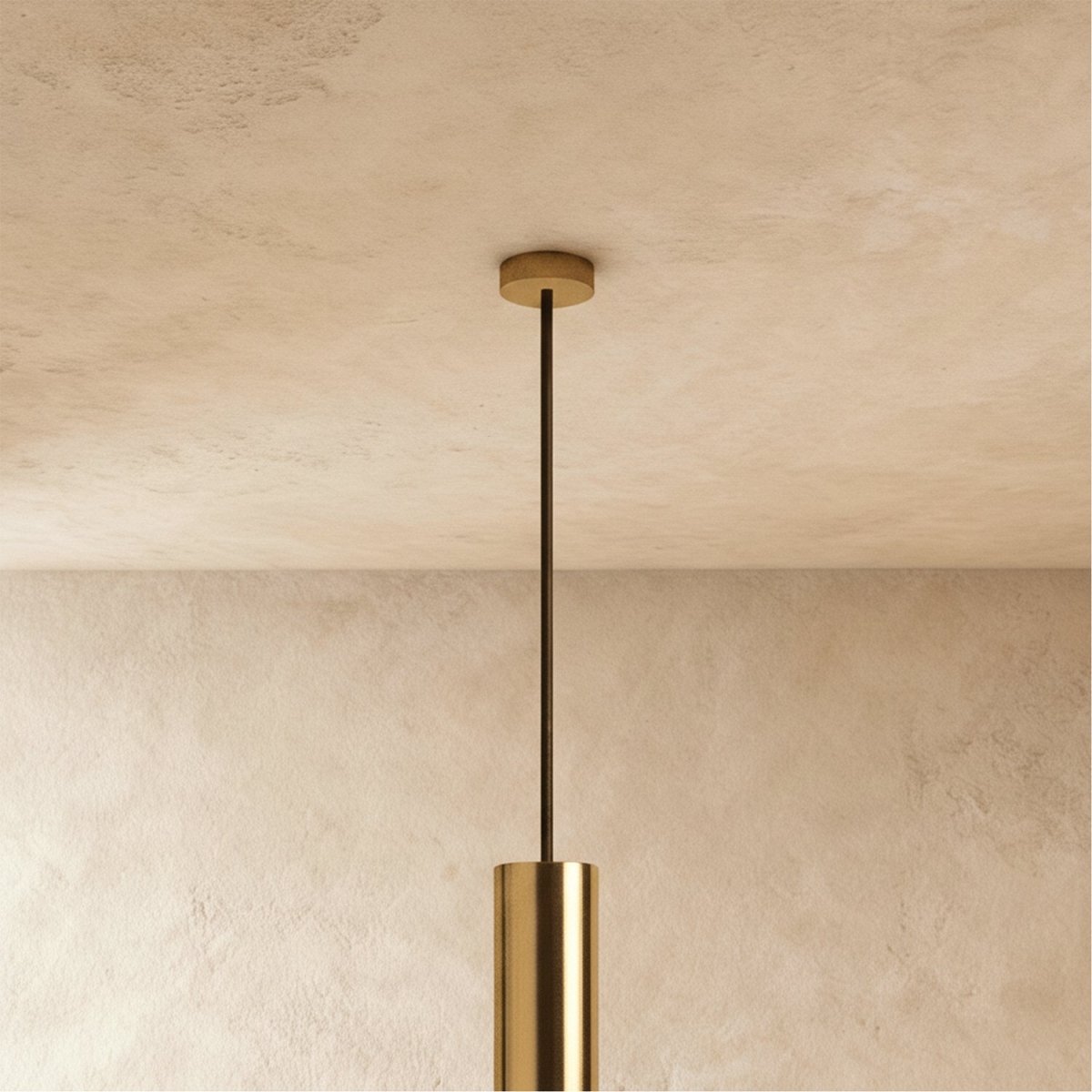 Clérachic gold-toned ceiling rose and slim dark rod leading down to the top of a gold-toned cylinder, against a plaster wall.