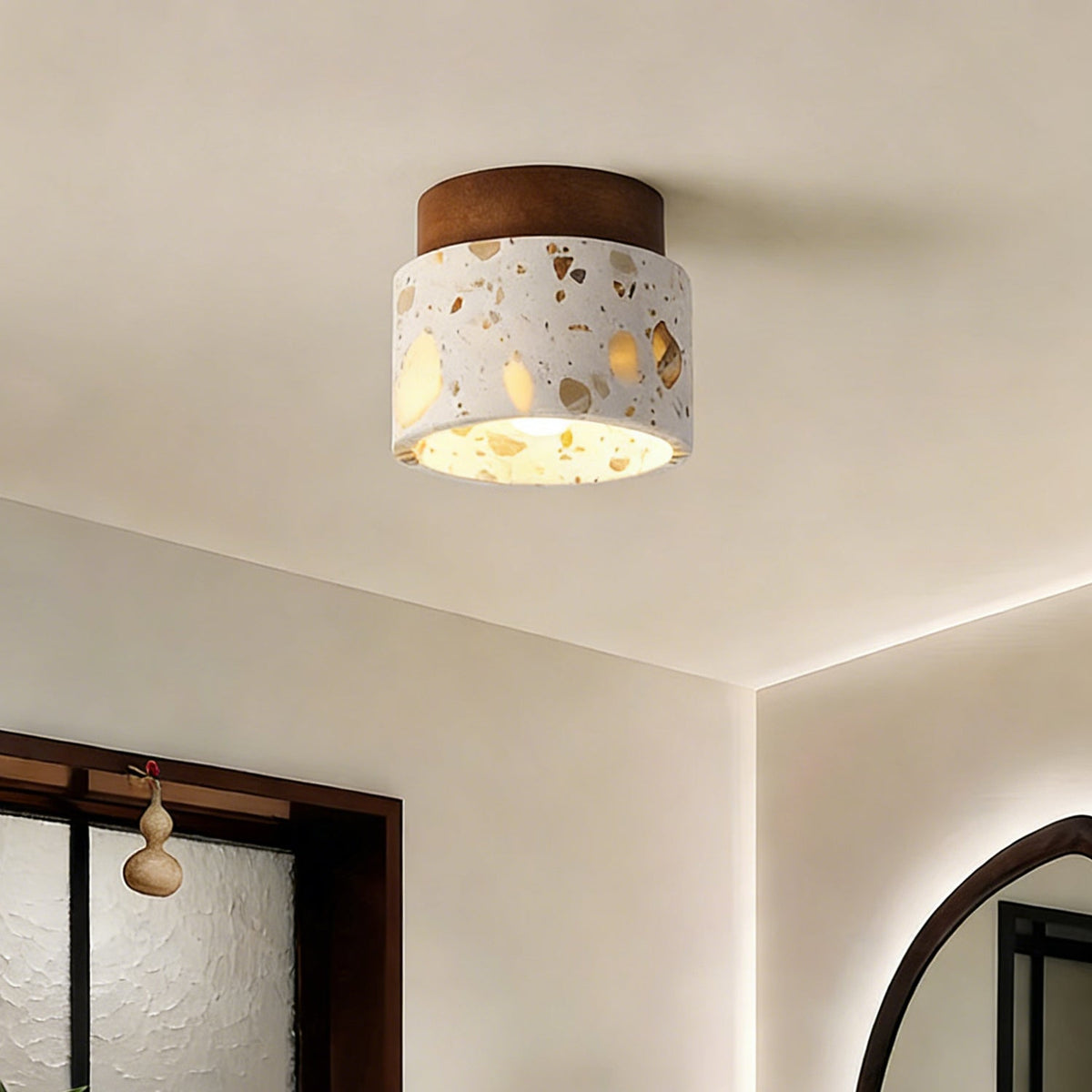 Terrazzo drum shade of this Clérachic ceiling light lit up near the corner of a cream room, with a round mirror and window beyond.