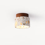 Compact Clérachic ceiling light with a terrazzo shade and walnut canopy, lit and viewed from slightly below on a pale backdrop.