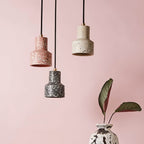 Against a soft pink wall beside a leafy plant, three Clérachic terrazzo pendants hang on black cords in pink, cream and charcoal.