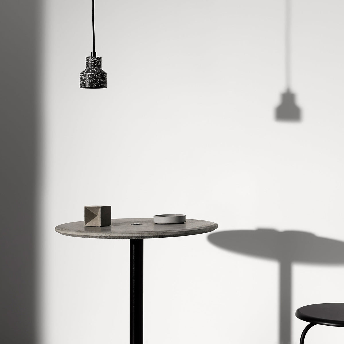 One small black-speckled Clérachic pendant hangs over a round table with a stool, casting a soft shadow on a pale wall.