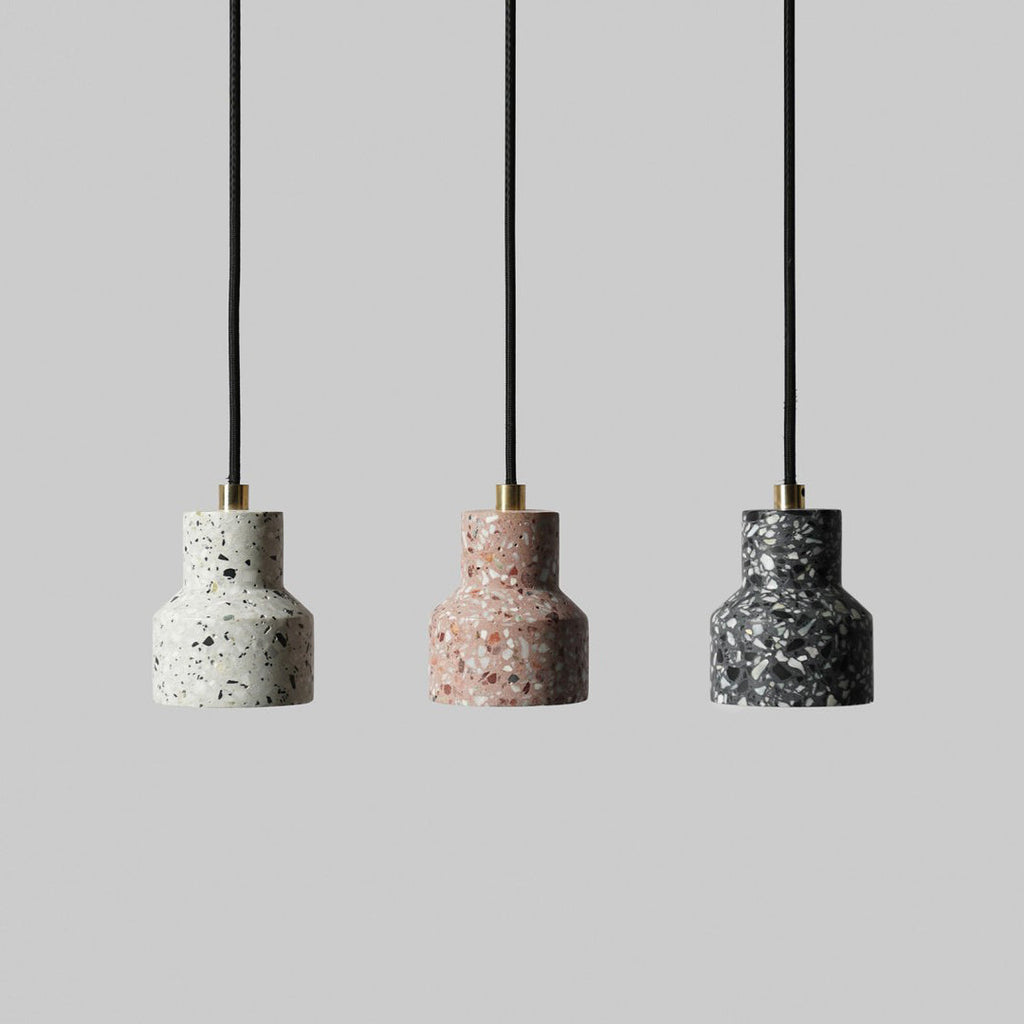 Three Clérachic terrazzo pendants on black fabric cords with brass tops, in white, rose and charcoal, against a soft grey backdrop.