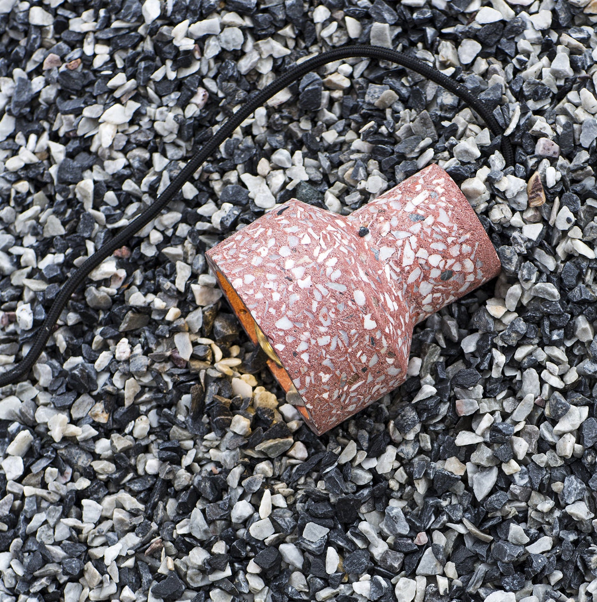 A rose-red terrazzo shade from Clérachic lies on its side on dark gravel, its black cord trailing and the warm inner lining showing.