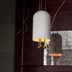 Close view of this Clérachic ribbed white pendant glowing gold inside its shade, with brass-toned balls below, against a dark wall with brass inlay lines.