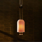 A Clérachic pendant lit to a warm peach glow, its ribbed shade hung from brass rods in front of a dark woven wall.