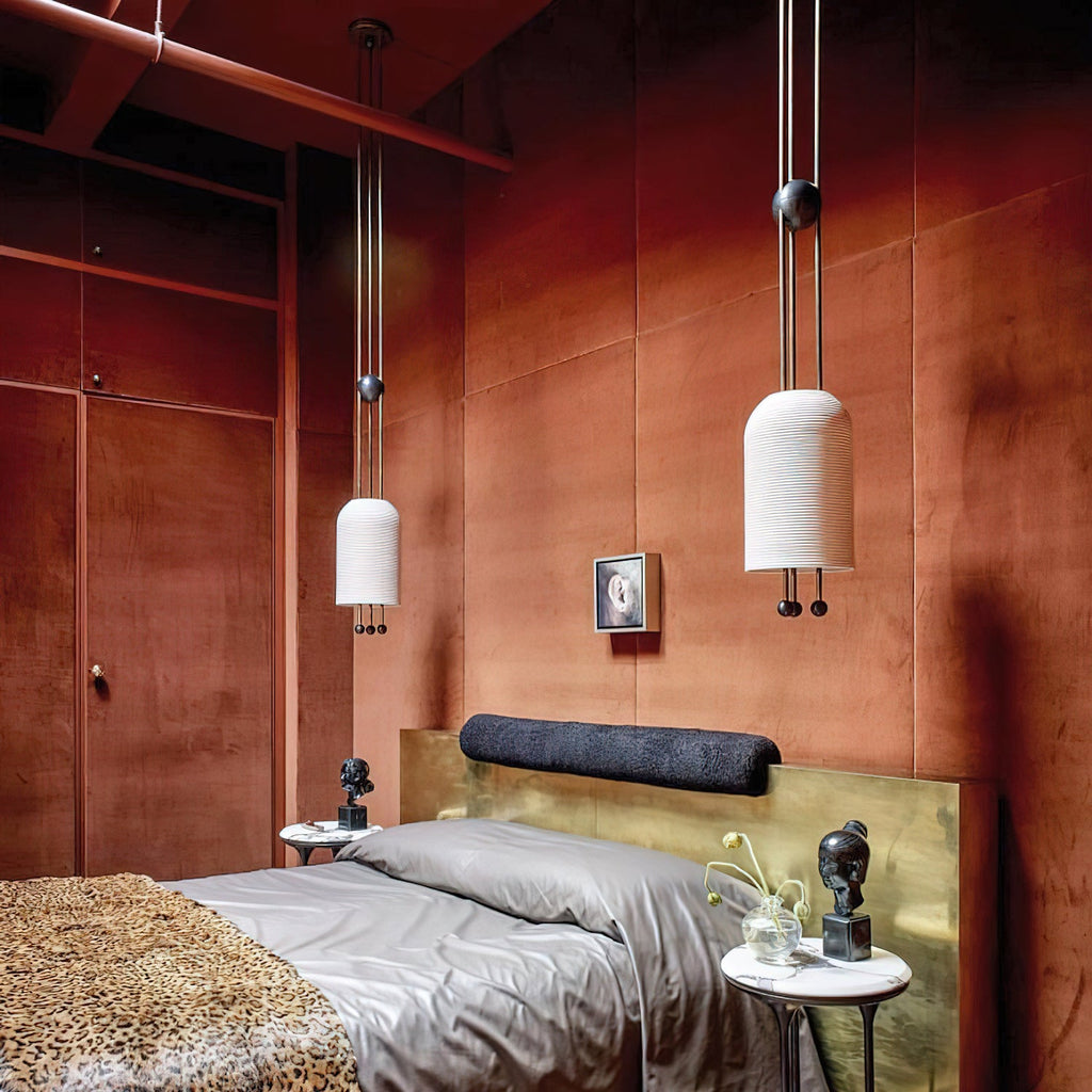 Two Clérachic pendants hung at different heights in a rust-red room, with a bed in grey linen and a leopard-print throw below.