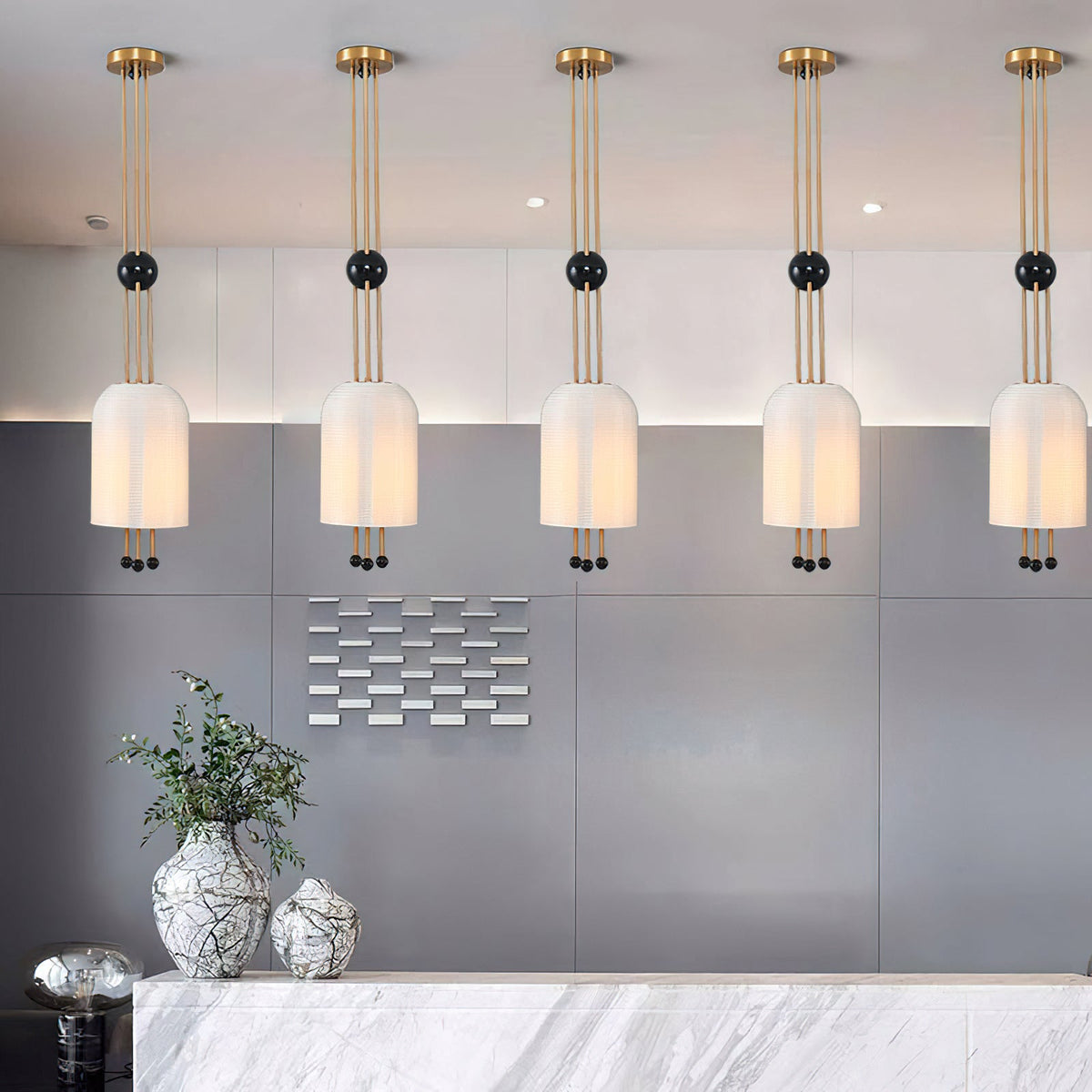 A row of five Clérachic white pendants hung from brass canopies above a pale marble counter, with a grey wall and a vase of foliage.