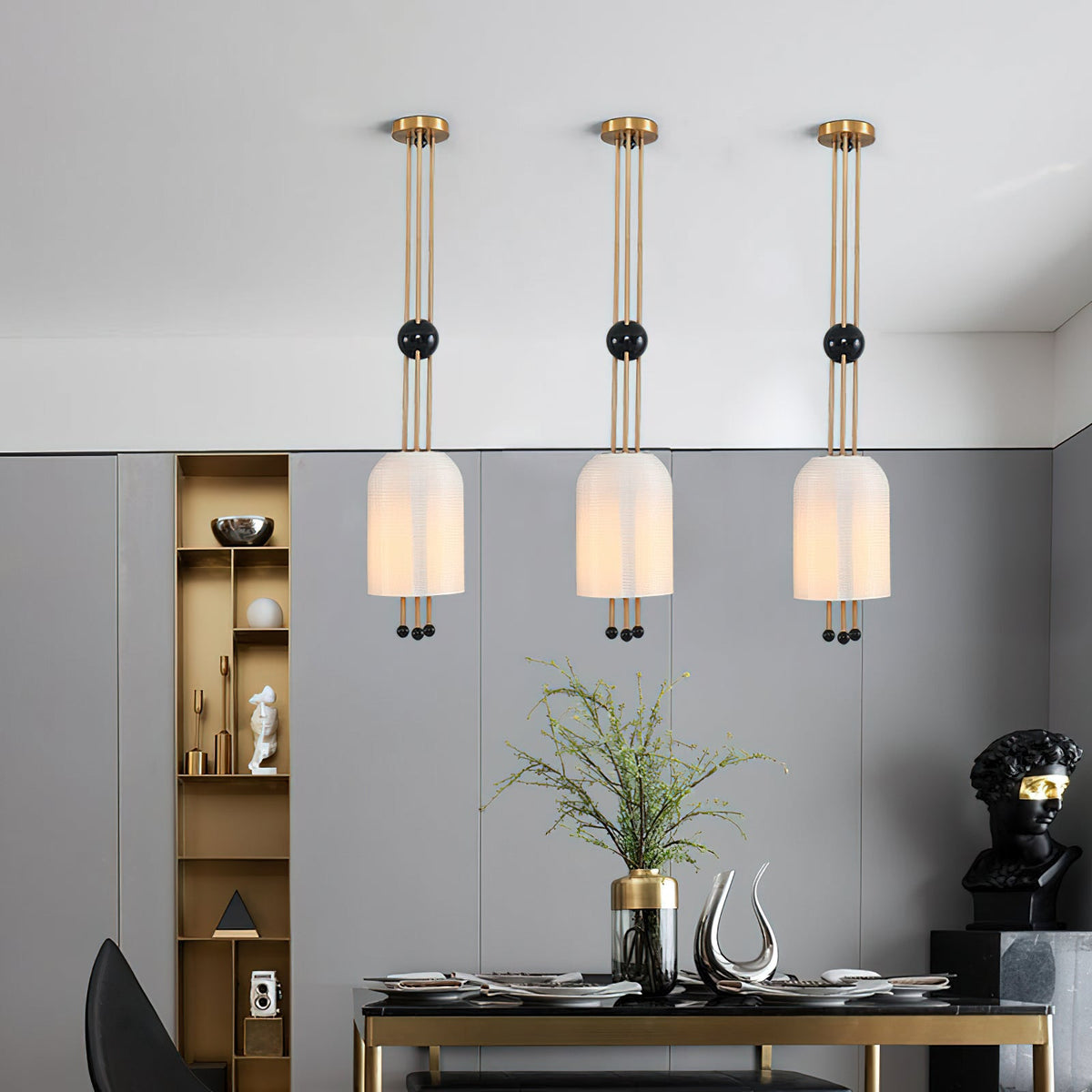 Three Clérachic pendants in a line above a dark dining table in a grey room with a brass shelving unit and a bust.