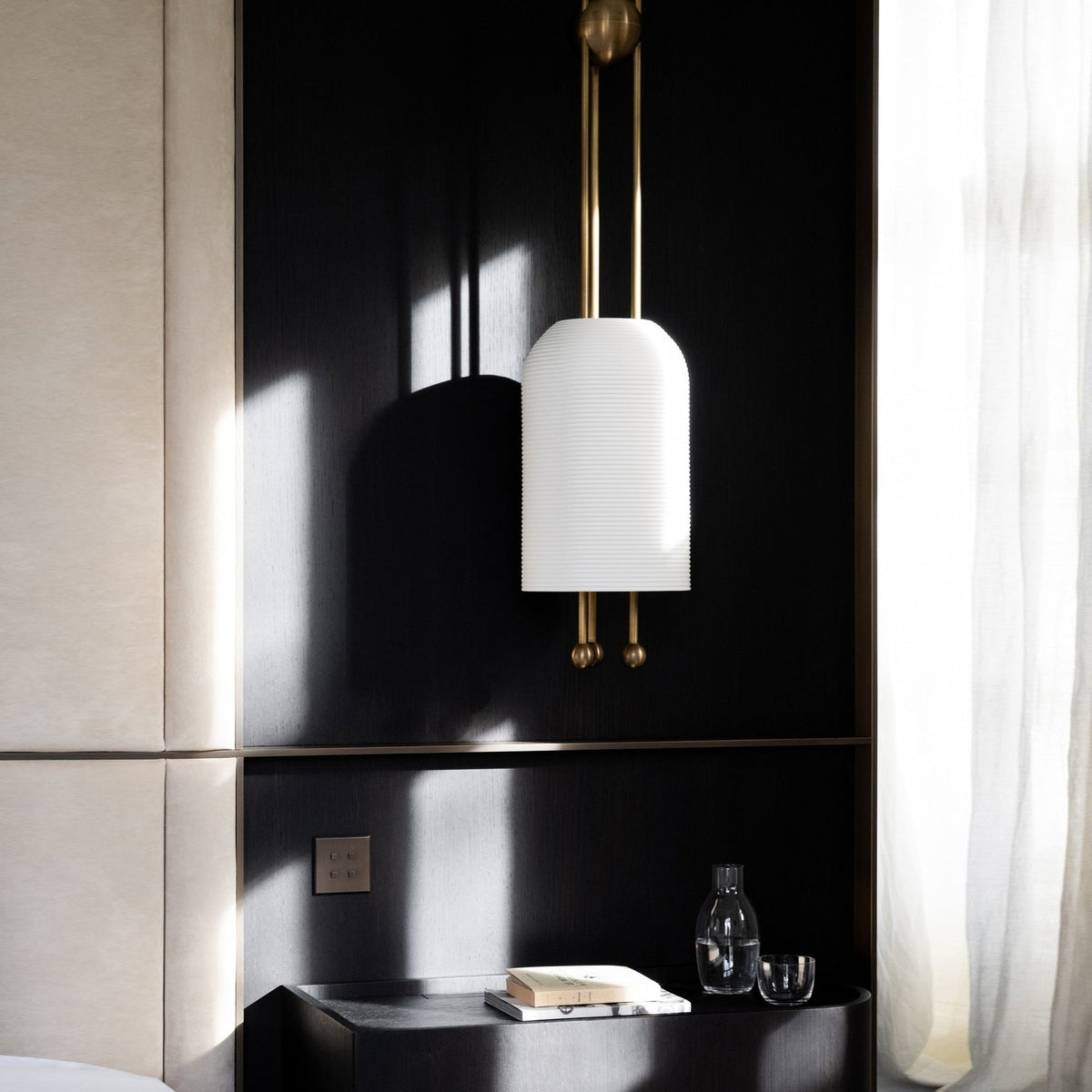 A single Clérachic white pendant on two brass rods against a black panelled wall, with a glass carafe and a book on a ledge below.