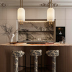 Two Clérachic pendants lit above a marble-backed kitchen island, with patterned stools and a branch in a white vase.