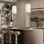 Two Clérachic pendants over a curved, fluted kitchen island with patterned stools, in front of a marble splashback and a palm mural.