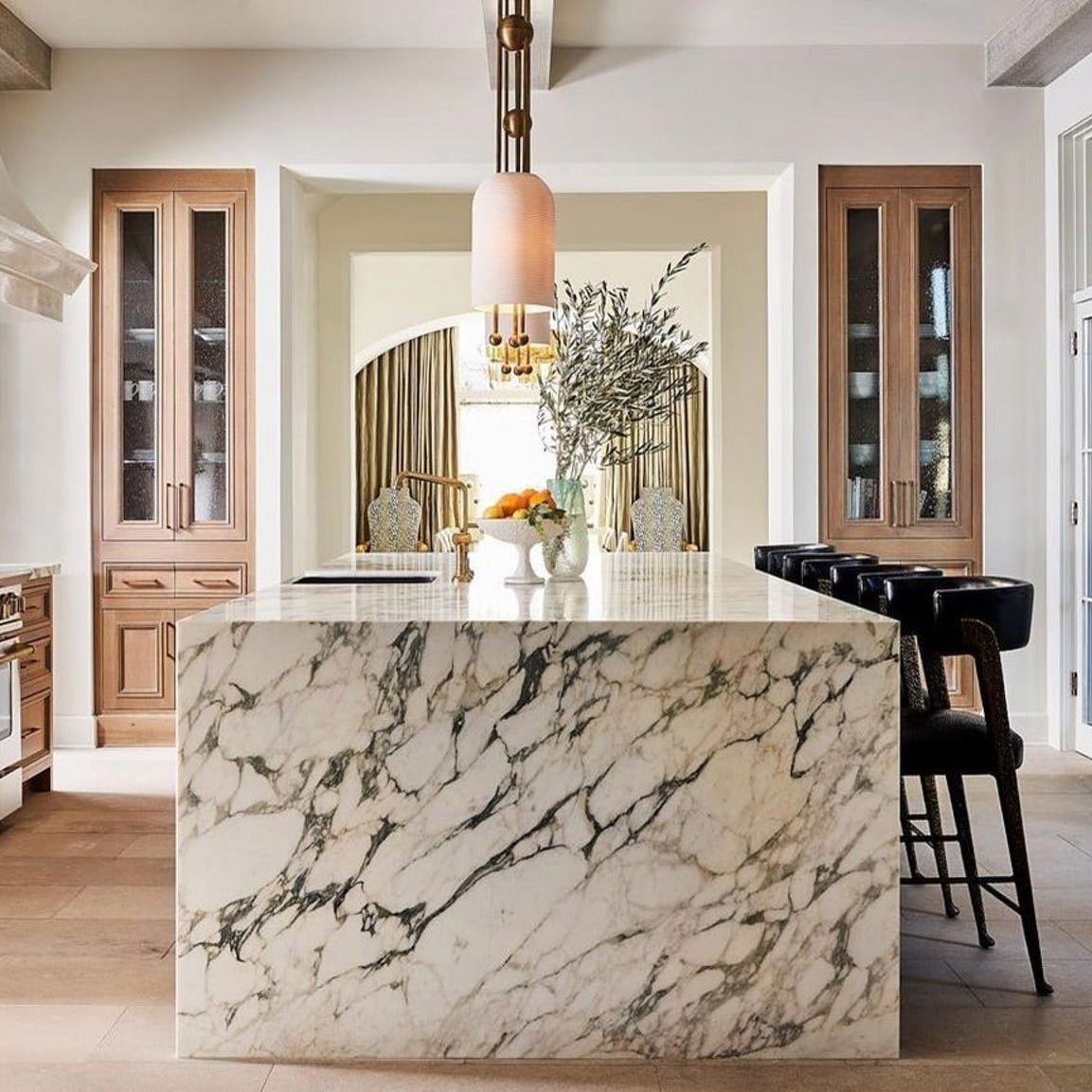 A Clérachic pendant with a warm-pink glow above a marble-clad kitchen island, with dark stools and tall wooden glass cabinets behind.