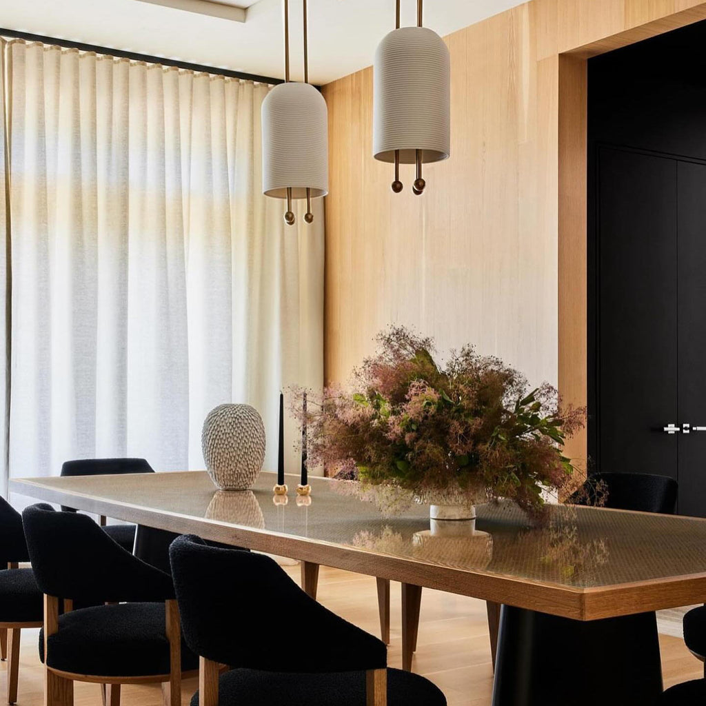 Two Clérachic pendants with pale stone-toned shades above a long dining table with black chairs and a bouquet, by a sheer curtain.