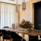 Two Clérachic pendants with pale stone-toned shades above a long dining table with black chairs and a bouquet, by a sheer curtain.