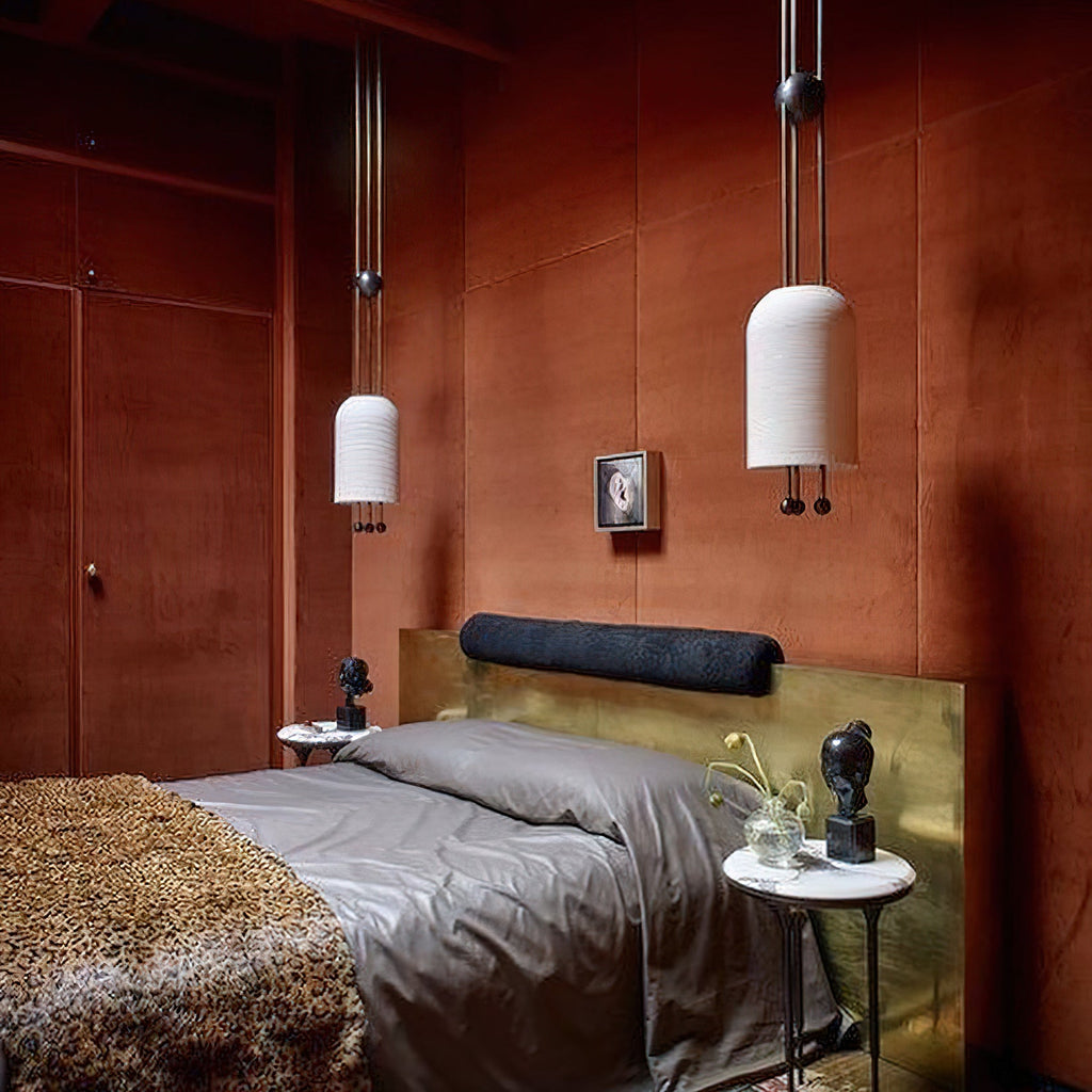A Clérachic pendant pair at different heights in a rust-toned bedroom, with a brass headboard, grey bedding and a small framed picture.