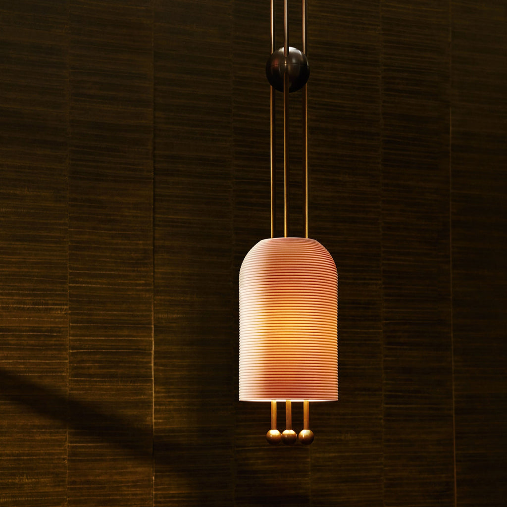 A Clérachic ribbed pendant glowing peach against a dark woven wall, with brass rods above and three brass balls below.
