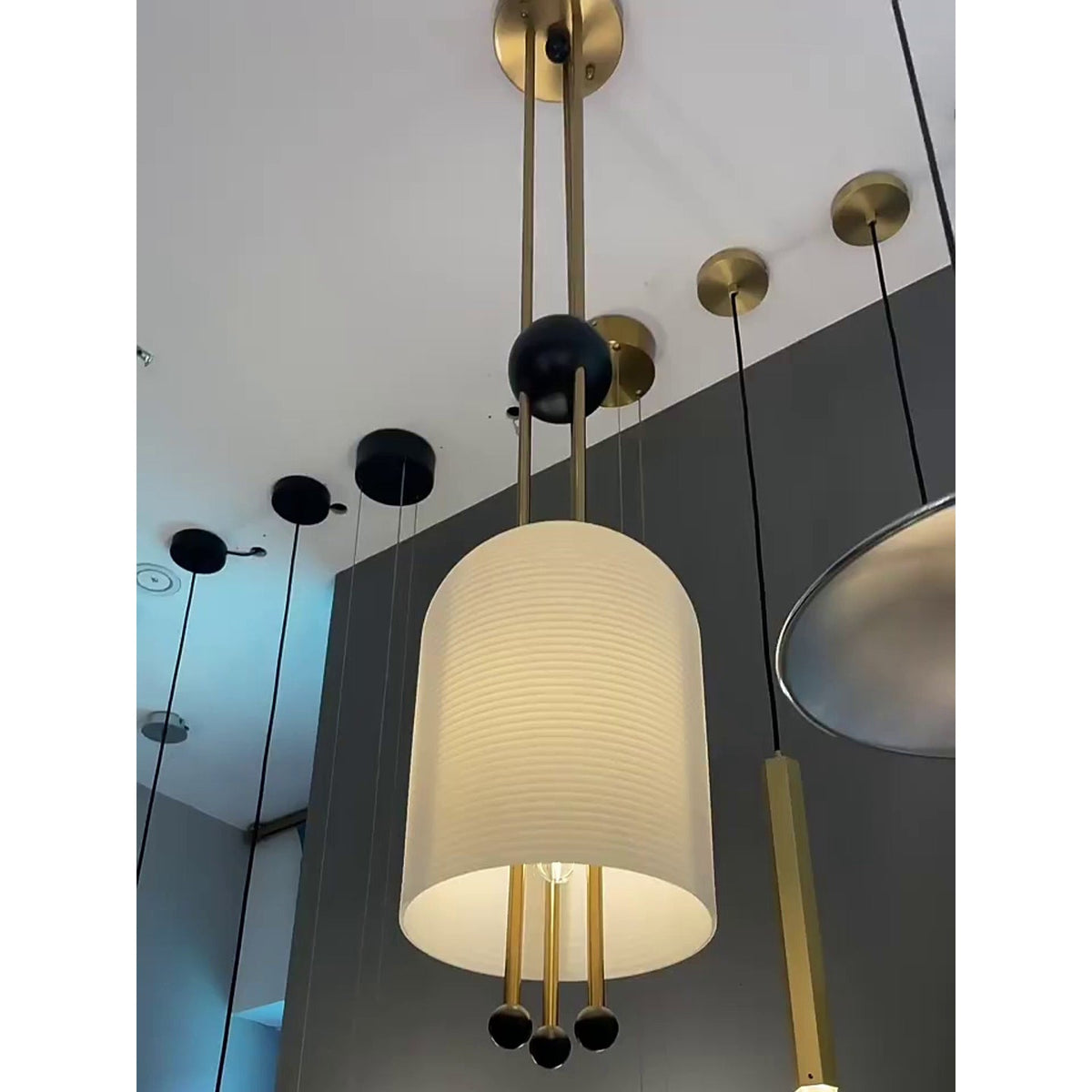 Looking up at this Clérachic pendant, its ribbed white shade lit and three brass-and-black balls below, in a showroom with other lights.