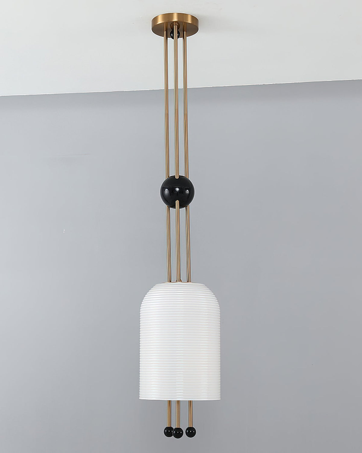A Clérachic ribbed white pendant against a grey wall, with its brass canopy at the ceiling and a black ball on the rods.