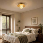 A Clérachic flush light with a frosted globe and bamboo disc sits above a bedroom with white bedding and a sage-green throw.