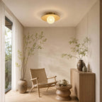 A white-globe Clérachic flush light with a bamboo disc hangs in a calm corner with a woven chair and a low wooden side table.