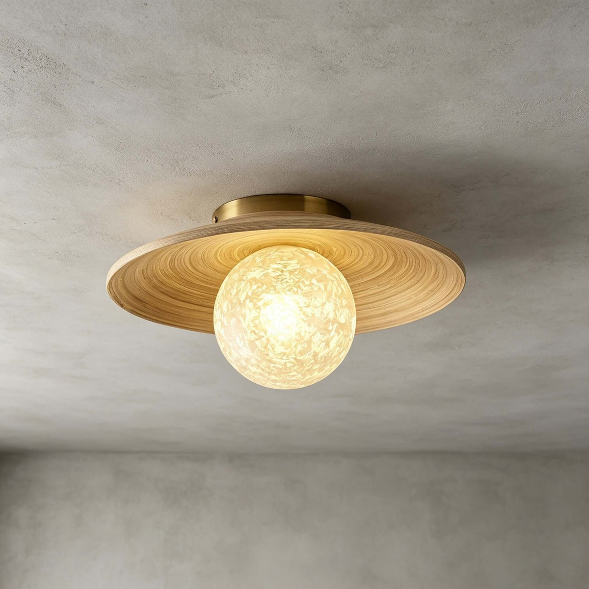 The white ice-textured glass globe and bamboo disc of this Clérachic ceiling light, with a brass canopy, on a grey wall.