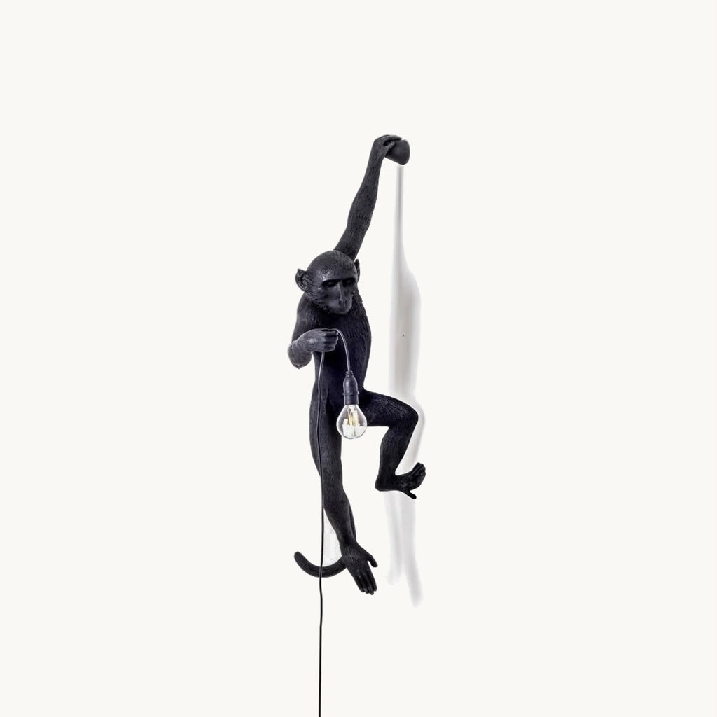 A black resin monkey wall light from Clérachic hangs by one arm on a plain white backdrop, holding a bare bulb on its cord.