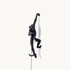 A black resin monkey wall light from Clérachic hangs by one arm on a plain white backdrop, holding a bare bulb on its cord.
