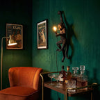 This Clérachic monkey wall light climbs a deep green wall beside an orange armchair and a drinks trolley, its bulb lit.