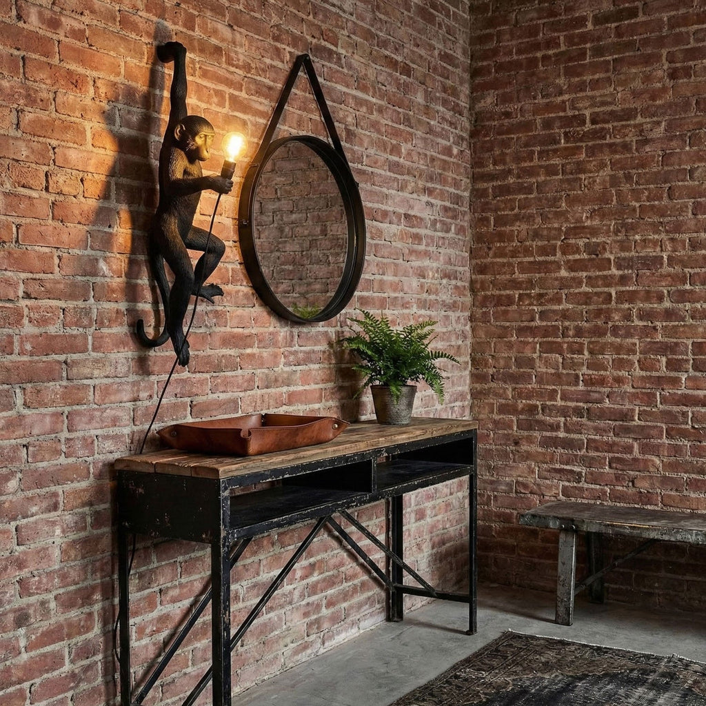 Against a red brick wall above a black metal console, a black Clérachic monkey light holds a glowing bulb beside a round mirror.
