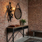 Against a red brick wall above a black metal console, a black Clérachic monkey light holds a glowing bulb beside a round mirror.