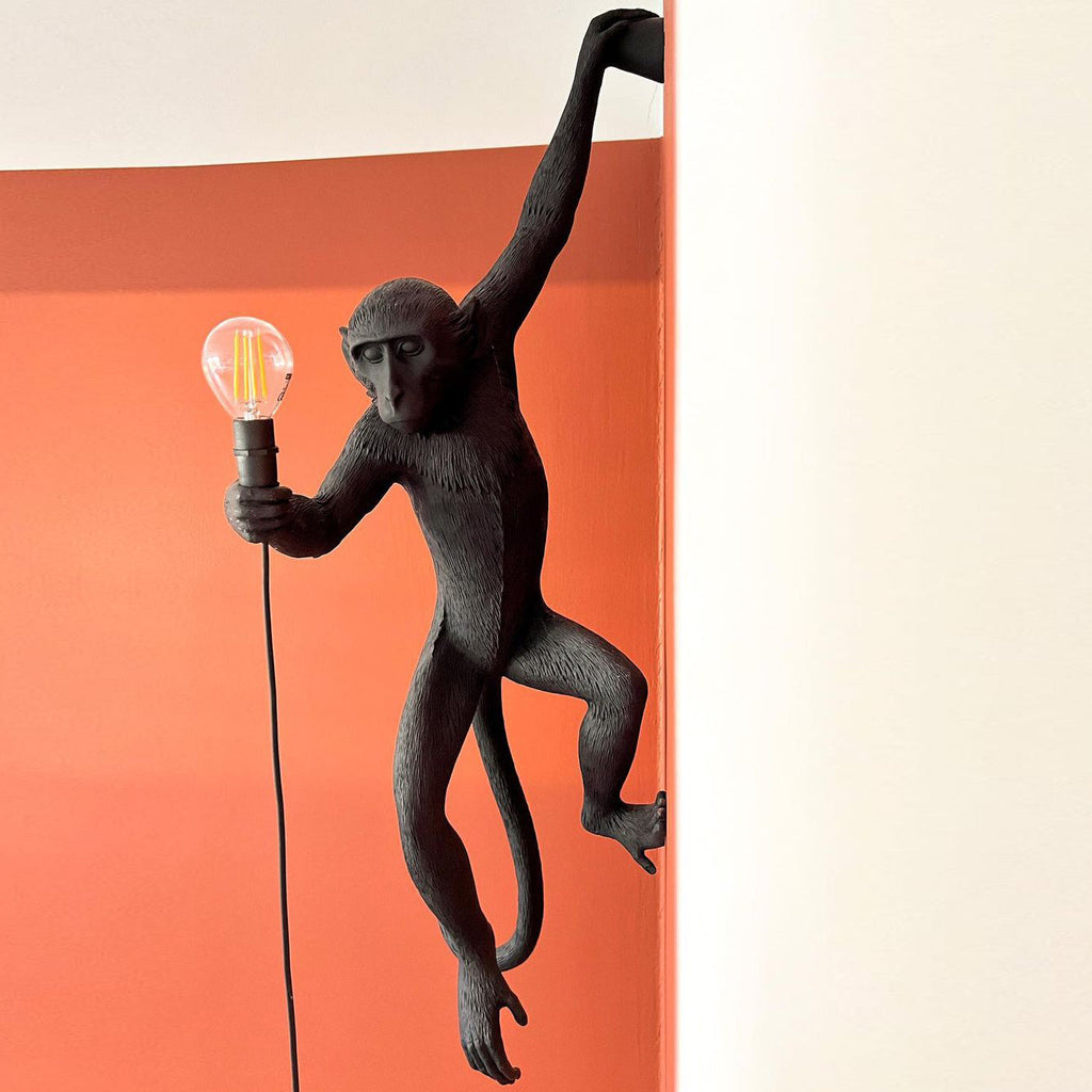 Close view of a black monkey on an orange wall, one arm raised, holding a bulb in a black socket on this Clérachic wall light.