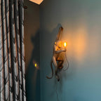 A gold-toned Clérachic monkey light clings to a teal-grey wall beside patterned curtains, its small bulb casting a warm glow.