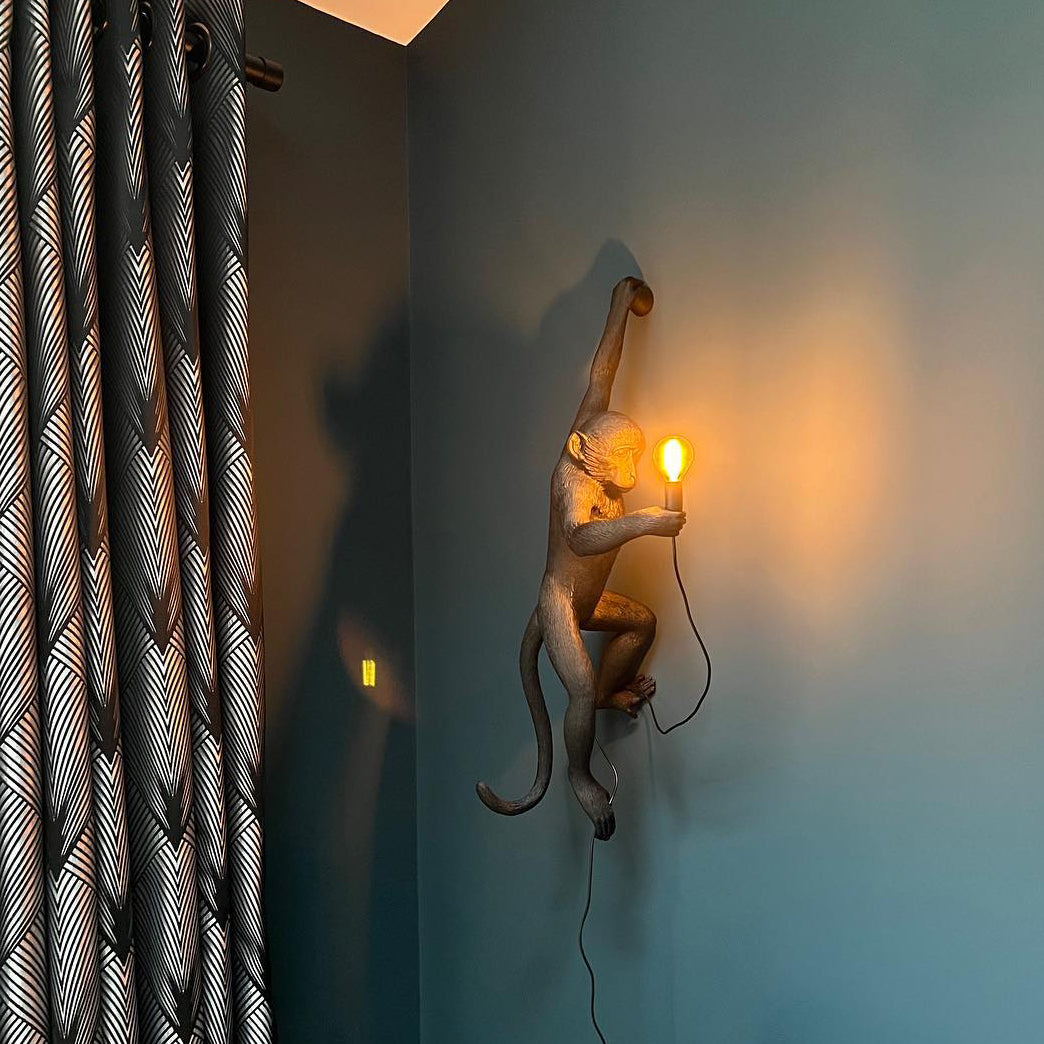 A gold-toned Clérachic monkey light clings to a teal-grey wall beside patterned curtains, its small bulb casting a warm glow.