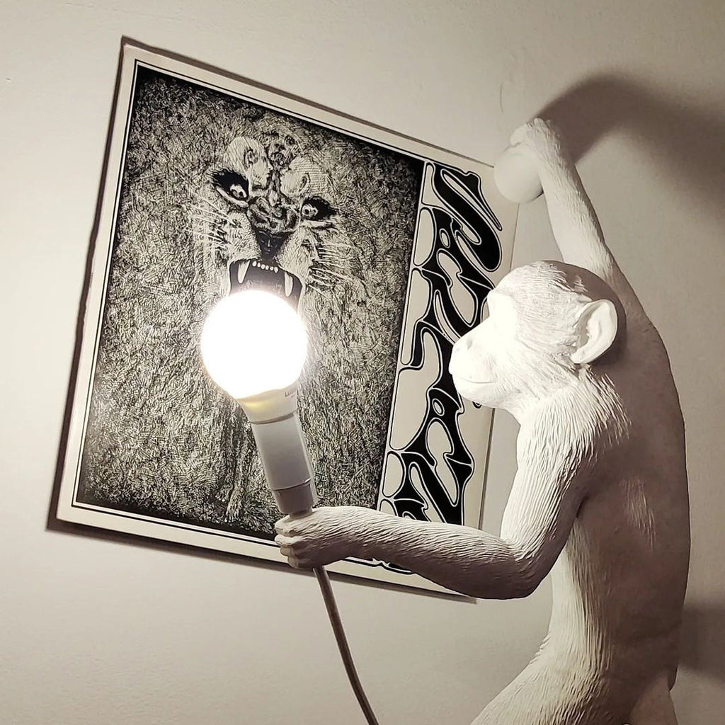 Close on the white resin monkey of this Clérachic wall light, a round bulb in its hand, set beside a large black-and-white poster.