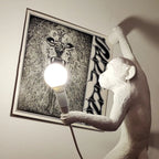 Close on the white resin monkey of this Clérachic wall light, a round bulb in its hand, set beside a large black-and-white poster.