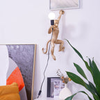 A gold Clérachic monkey wall light holds a white globe bulb above a pale cabinet, with a large green plant at the right.