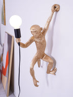 Gold monkey of this Clérachic wall light seen close up, one arm gripping the wall and the other holding a black socket and bulb.