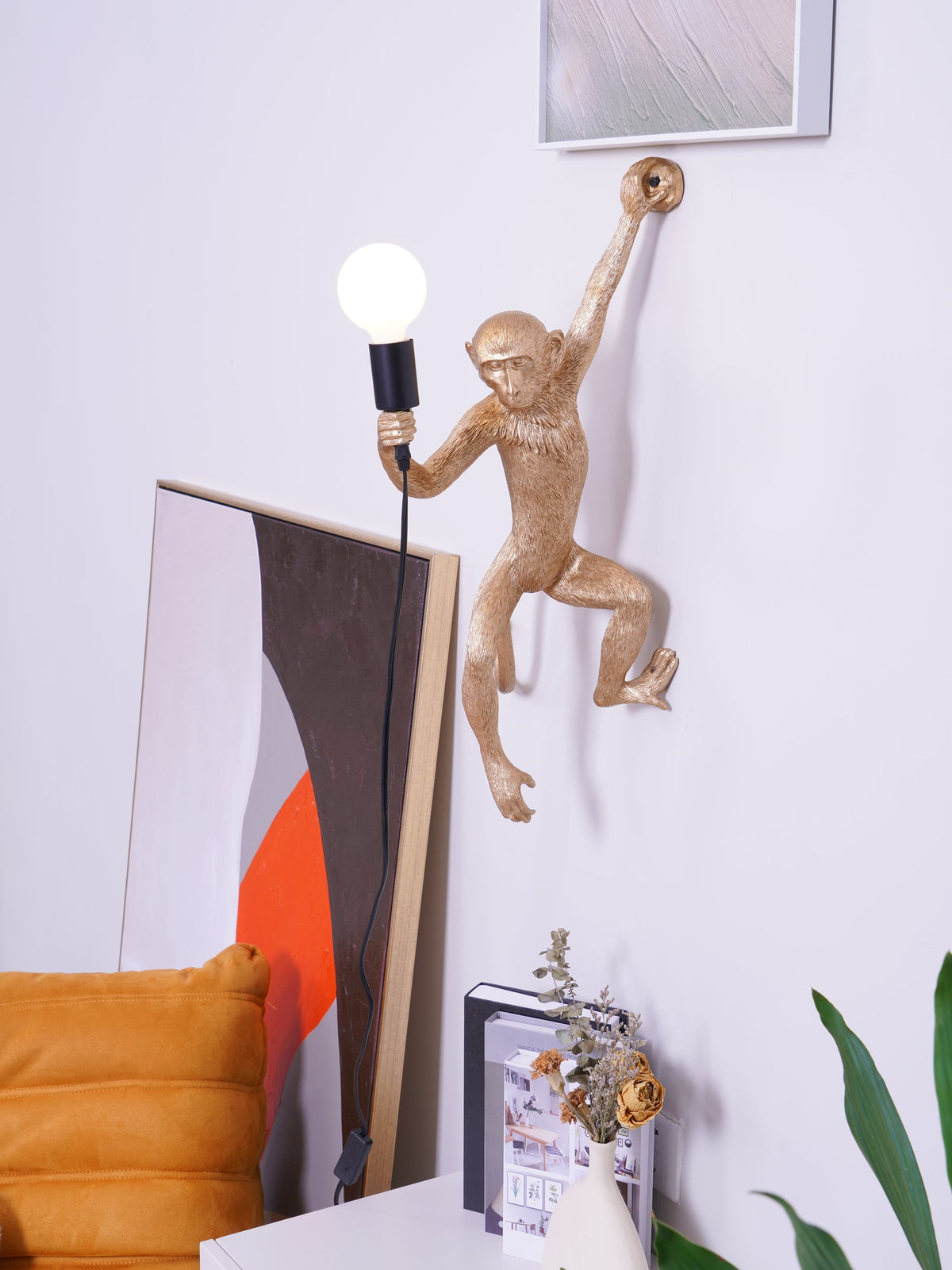 A gold monkey wall light from Clérachic reaches up to the wall with a bulb in hand, framed art and a cream vase below.