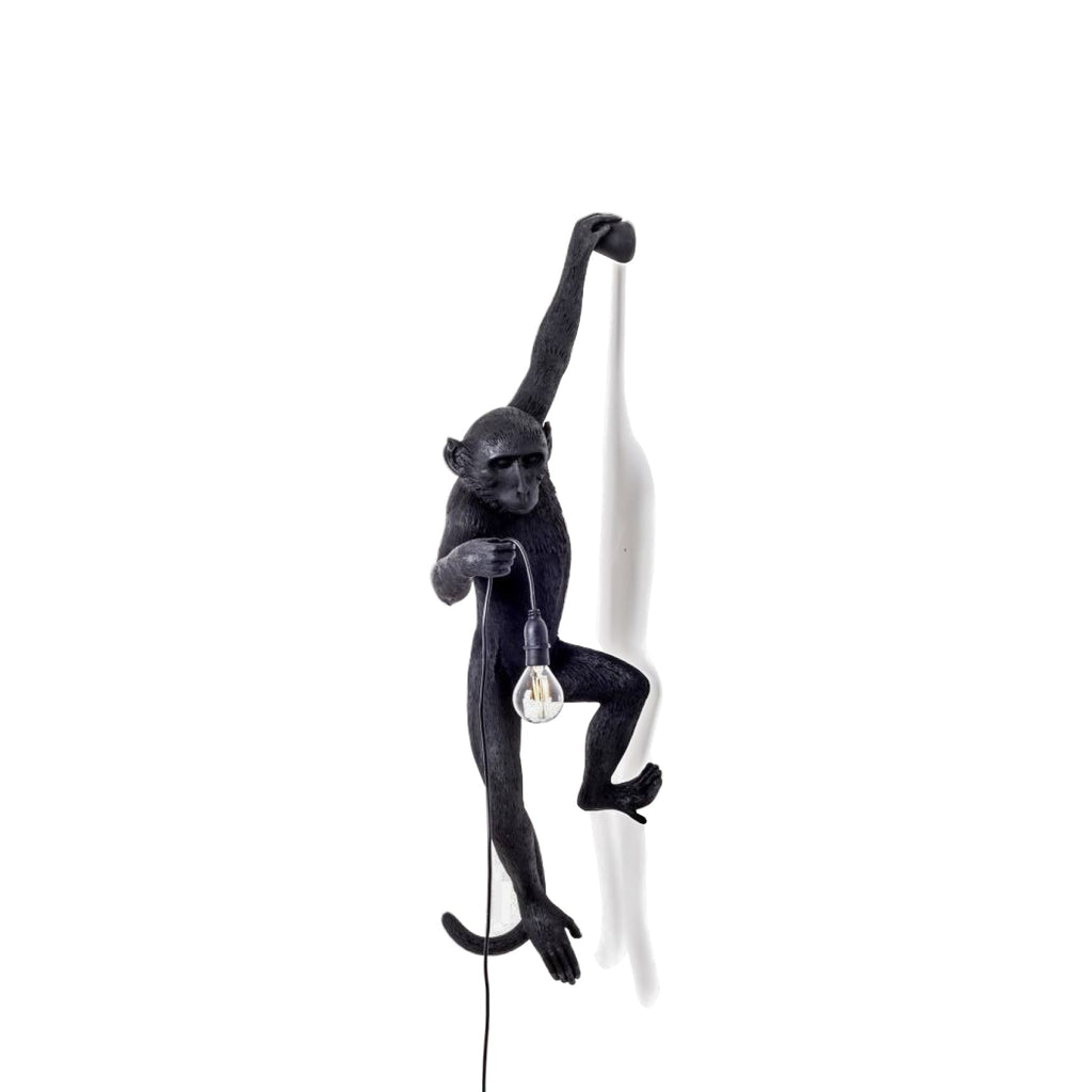 The black monkey of this Clérachic wall light hangs from one arm over a white backdrop, holding a filament bulb at waist height.