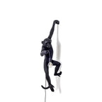 The black monkey of this Clérachic wall light hangs from one arm over a white backdrop, holding a filament bulb at waist height.