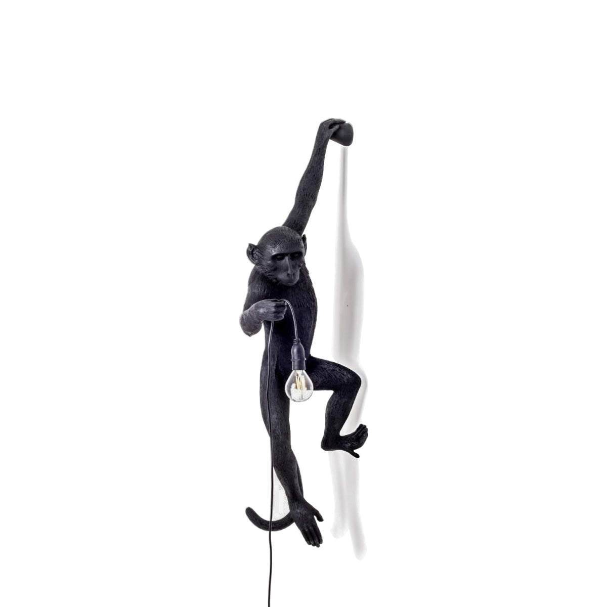The black monkey of this Clérachic wall light hangs from one arm over a white backdrop, holding a filament bulb at waist height.