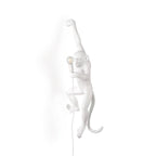 A white resin monkey wall light from Clérachic hangs by one arm on a white background, its bulb held close to its body.